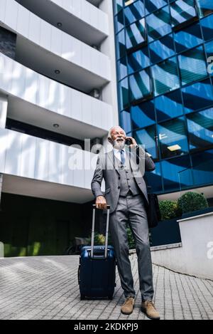 Confident smiling entrepreneur talking on smart phone. Senior businessman wearing suit is carrying laptop bag and holding suitcase. He is standing aga Stock Photo