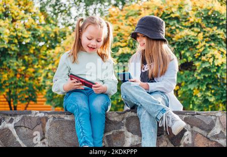 Two smiling little sister girls kids sitting and browsing their smartphone devices in the autumnal park. Careless young childhood time and a modern te Stock Photo