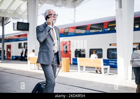 Senior businessman talking on smart phone while walking with luggage. Male professional is in suit at subway station platform. He is commuting in the Stock Photo