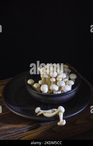 Fresh uncooked bunapi white shimeji edible mushrooms from Asia in a ceramic bowl on a dark wooden background. Stock Photo