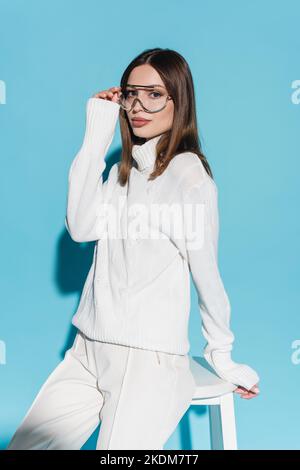 young and stylish woman in white outfit adjusting eyeglasses while leaning on high chair on blue Stock Photo