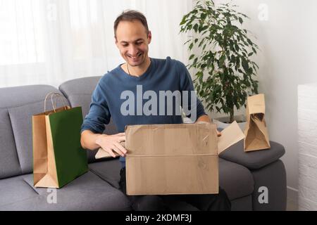 Present from distant friend. Happy millennial man unpack open box with birthday gift surprise received by mail. Smiling young guy has pleasure to Stock Photo