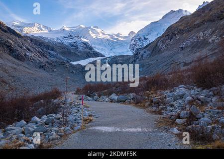 Hiking path in the shade to the Morteratsch glacier in Switzerland. Above the glacier, the mountains still light up brightly by the low sun. Stock Photo