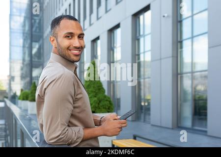 Portrait of African American businessman, man holding phone smiling and looking at camera, office worker in shirt outside modern office building on lunch break. Stock Photo