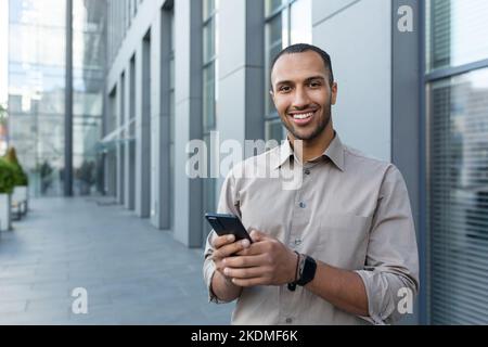 Portrait of African American businessman, man holding phone smiling and looking at camera, office worker in shirt outside modern office building on lunch break. Stock Photo