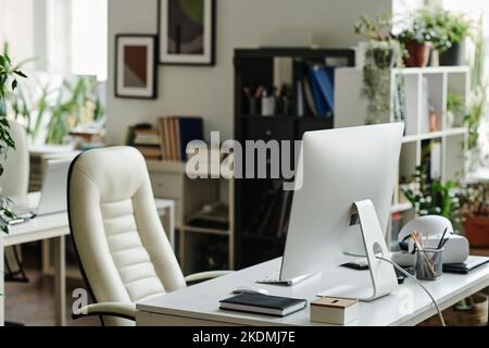 Computer monitor standing on workplace of office worker with notebook and other supplies and white leather armchair near by Stock Photo