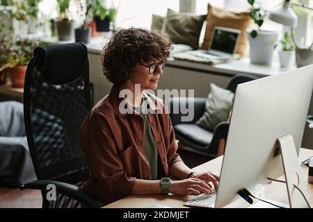 Young serious businesswoman typing on keyboard in front of computer monitor while working over new website in openspace office Stock Photo