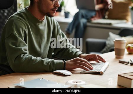 Close-up of young businessman in sweatshirt typing on computer keyboard while sitting by workplace against female colleague Stock Photo