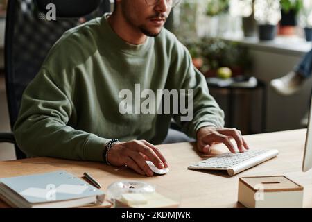 Young man in casualwear clicking mouse and pressing key of computer keyboard while working over new software by workplace Stock Photo