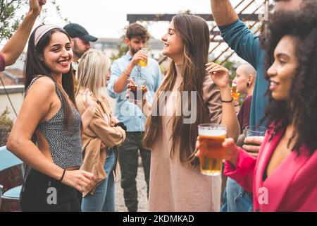 Happy community of multiethnic friends having fun dancing together at rooftop party - young people cheering drinking beers in the terrace - internatio Stock Photo