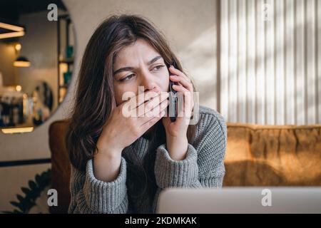 Sad young woman talking on the phone while sitting in front of a laptop. Stock Photo