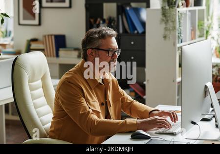 Serious mature businessman or chief executive officer sitting by workplace in front of computer monitor and networking Stock Photo