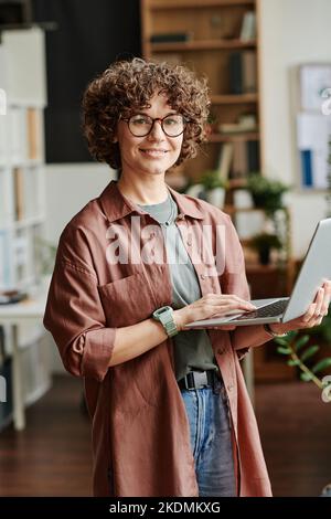 Young successful female designer with laptop standing in office and looking at camera while searching for online data or video Stock Photo