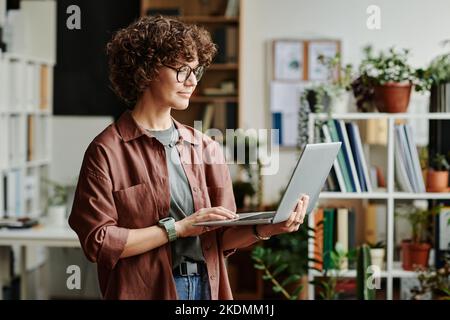 Young confident brunette female programmer with laptop looking at screen while reading online information or watching video Stock Photo