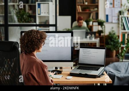 Rear view of young brunette female IT specialist sitting by desk in front of computers with coded data while trying to decode it Stock Photo