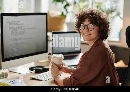 Young successful IT specialist with cup of coffee looking at camera while sitting by workplace with computer monitor and laptop Stock Photo