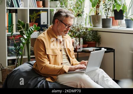 Serious mature businessman in casualwear and eyeglasses with laptop on knees networking in office while sitting on soft bag chair Stock Photo