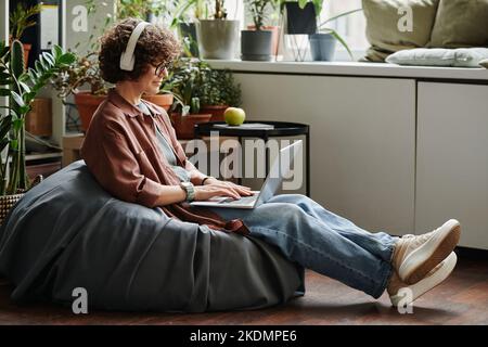 Young female employee in headphones and casualwear listening to music and networking while sitting in soft bag chair in modern office Stock Photo