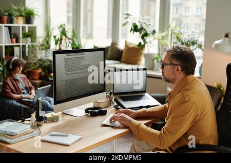 Serious businessman concentrating on decoding data while sitting by workplace in front of computer monitor and laptop in office Stock Photo