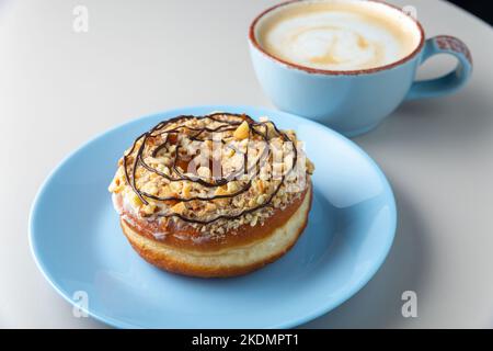 Donut topped with peanut sprinkles and chocolate topping on blue plate and cup of cappuccino with milk foam on a white table, normal view. Copy space Stock Photo