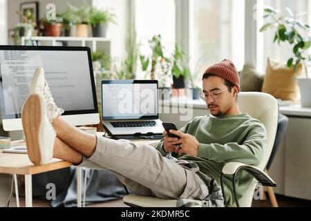 Young serious man in casualwear watching online video in smartphone or texting while sitting in armchair by his workplace in office Stock Photo