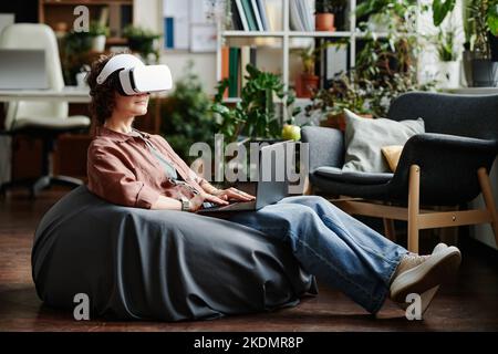 Young modern businesswoman in vr headset and laptop on her knees sitting on bag chair and preparing presentation of new project Stock Photo