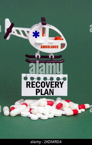 Medical concept. On a green surface, an ambulance helicopter, pills and a white sign with the inscription - RECOVERY PLAN Stock Photo