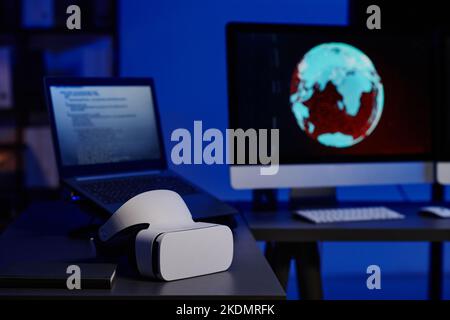 Selective focus on VR headset lying on workplace of programmers with laptop and computer monitor in dark room or openspace office Stock Photo