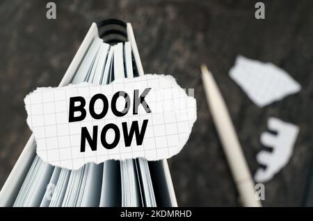 On the table is a notebook on which lies a piece of torn paper with the inscription - book now. The pen lies outside the sharpness zone. Stock Photo