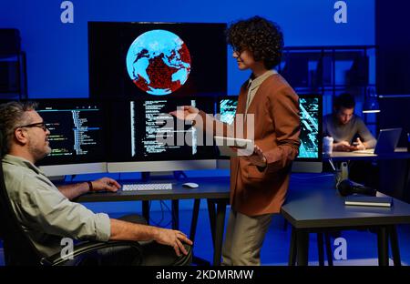 Young confident female programmer making report to mature male colleague while pointing at computer screen with coded data Stock Photo