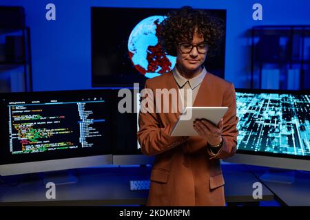 Young serious female programmer using tablet while standing against computer monitors with coded data on screens in dark room Stock Photo