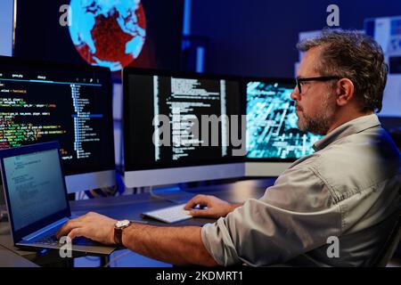 Side view of serious server engineer using laptop and computers while sitting in front of monitors by workplace and decoding data Stock Photo