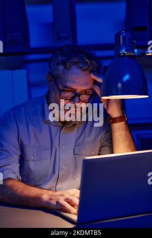 Tired mature male programmer sitting by workplace in front of laptop in office and finding ways of decoding data while working overtime Stock Photo