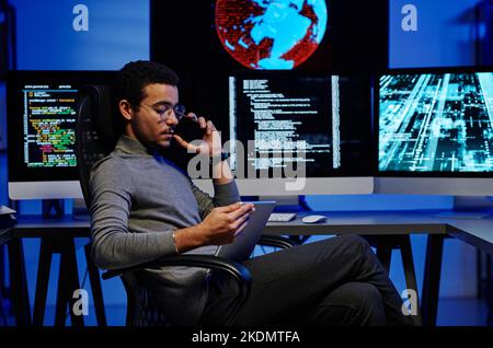 Side view of young busy male programmer talking on smartphone and looking through online information or video on tablet screen Stock Photo