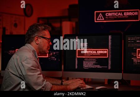 Side view of serious mature IT support manager trying to solve technical problem while sitting in front of computer screens with critical errors on Stock Photo