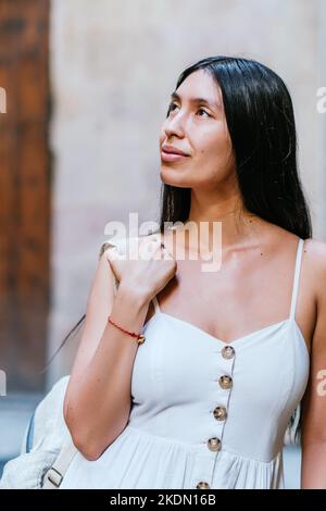 Dreamy Hispanic woman in white dress with backpack looking away and admiring historic street in Barcelona, Spain Stock Photo