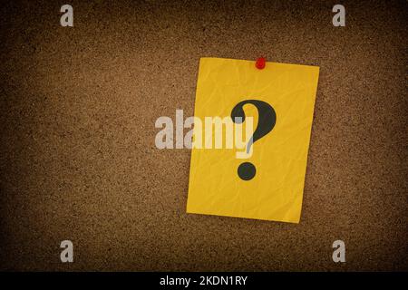 Yellow crumpled paper note with question marks on it pinned to a cork board. Closeup. Stock Photo