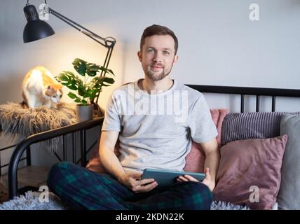 Attractive bearded male freelance software engineer sitting at home working using tablet with tabby cat sitting nearby. Young handsome developer man in gray t-shirt. High quality image Stock Photo