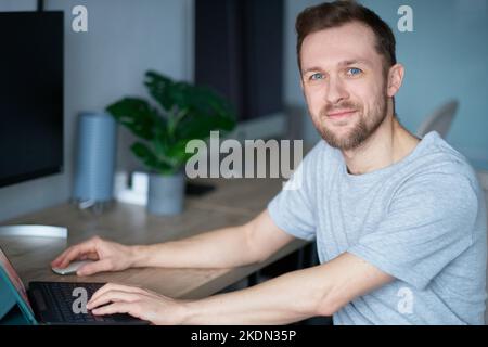 Attractive bearded male software engineer or web designer sitting at working place in home office hardly working using laptop. Young handsome man in grey t-shirt, positive emotion with computer Stock Photo
