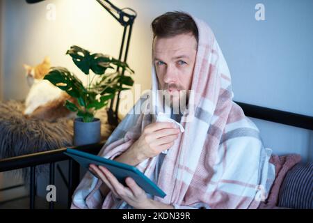 Feeling sick or illness concept. Bearded young man sneezing and using tablet in bed full covered with blanket and white ginger cat near him. Work from home office or online studying Stock Photo