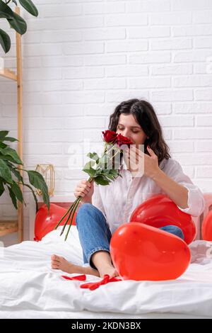 girl in love with bouquet of rose flowers on bed red heart shape balloons. Woman rejoices gift on valentines day. Easy-going girl with long hair Stock Photo