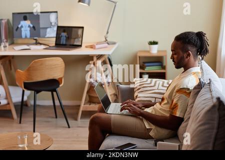 Side view portrait of creative black man using laptop while sitting on sofa in modern home interior, copy space Stock Photo