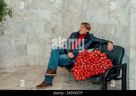 handsome man with a huge bouquet of scarlet roses is sitting in the waiting room. A happy man congratulates his wife on the birth of a child, on a wed Stock Photo