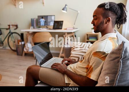 Side view portrait of creative black man using laptop while sitting on sofa and smiling at blank screen Stock Photo