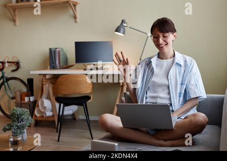 Portrait of young woman waving for video chat and smiling while using laptop on sofa with legs crossed Stock Photo