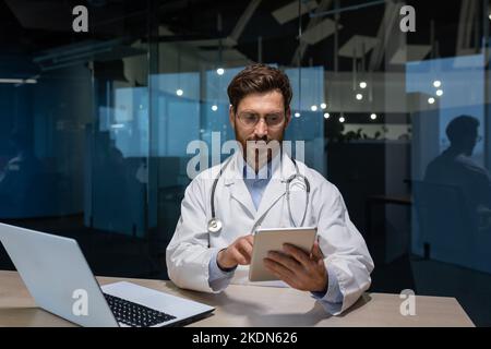 mature doctor in a medical gown is working inside clinic office, doctor is holding a tablet computer in his hands for online consultation of patients, working with a laptop. Stock Photo