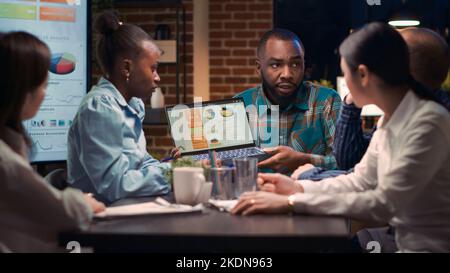 Business meeting, african american analytics specialist showing company revenue report data on laptop screen. Coworkers discussing statistics presentation, employees listening to analyst speaking Stock Photo