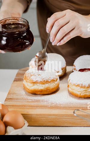 Cooking traditional Hanukkah sufganiyot. A woman decorates donuts with jam. Festive Jewish dessert. Stock Photo