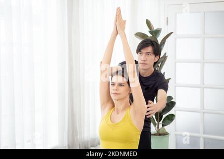 Young coach man training woman practicing yoga while stretching hands and balance in the room at home, instructor assistance female training workout y Stock Photo