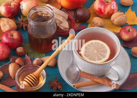 White table with autumn decorations, a cup of tea, cinnamon pieces, a glass bowl with honey, small apples, nuts, colourful tree leaves and acorns. Hig Stock Photo
